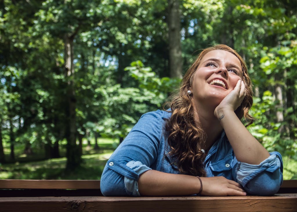 A smiling woman in a park leaning on a wooden railing, surrounded by lush foliage.