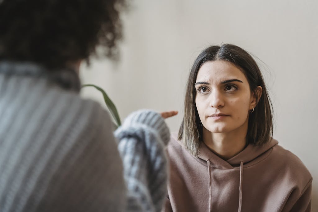 Sad young ladies in casual clothes standing in light apartment near wall while having disagreement and pointing finger in face while looking at each other